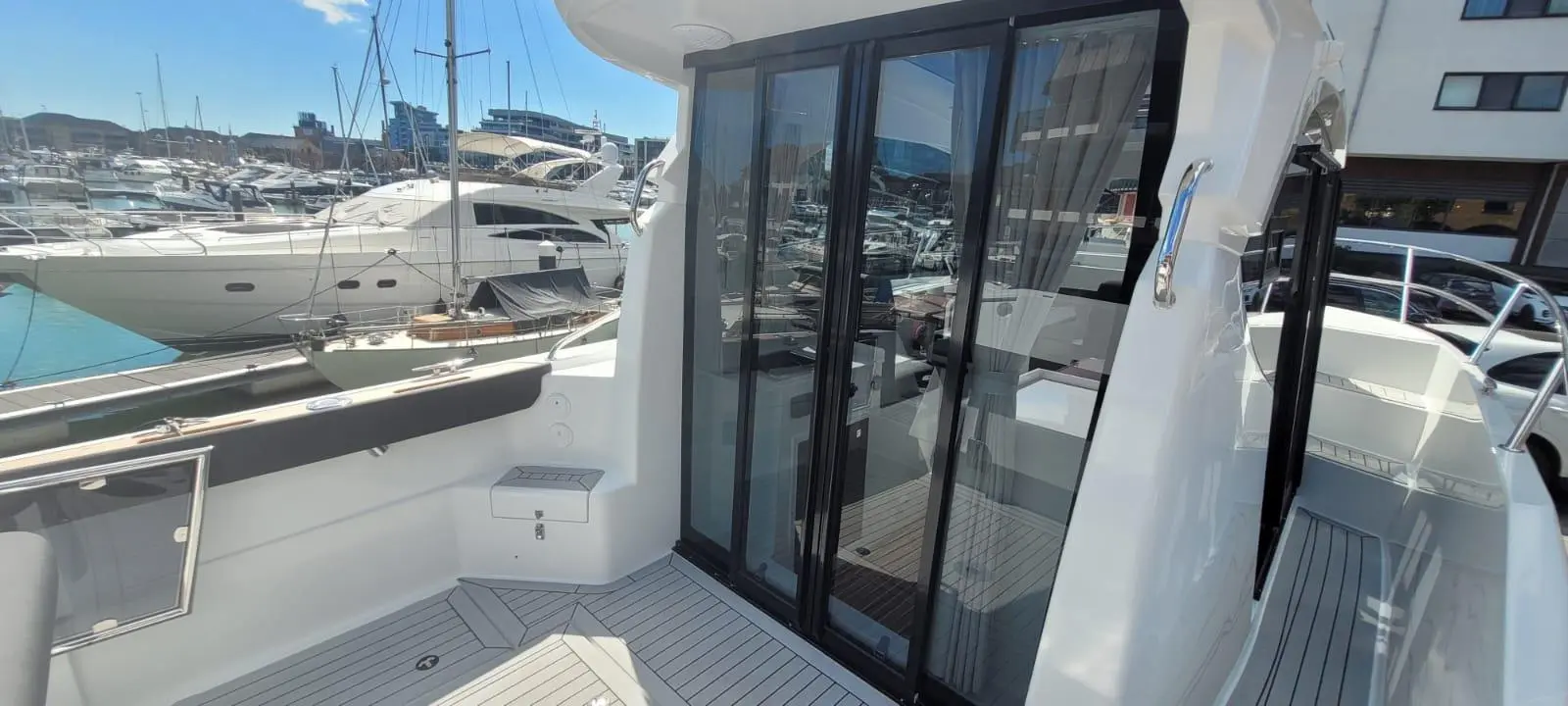 View of a sleek boat deck with glass doors, overlooking a marina filled with yachts under a clear blue sky.